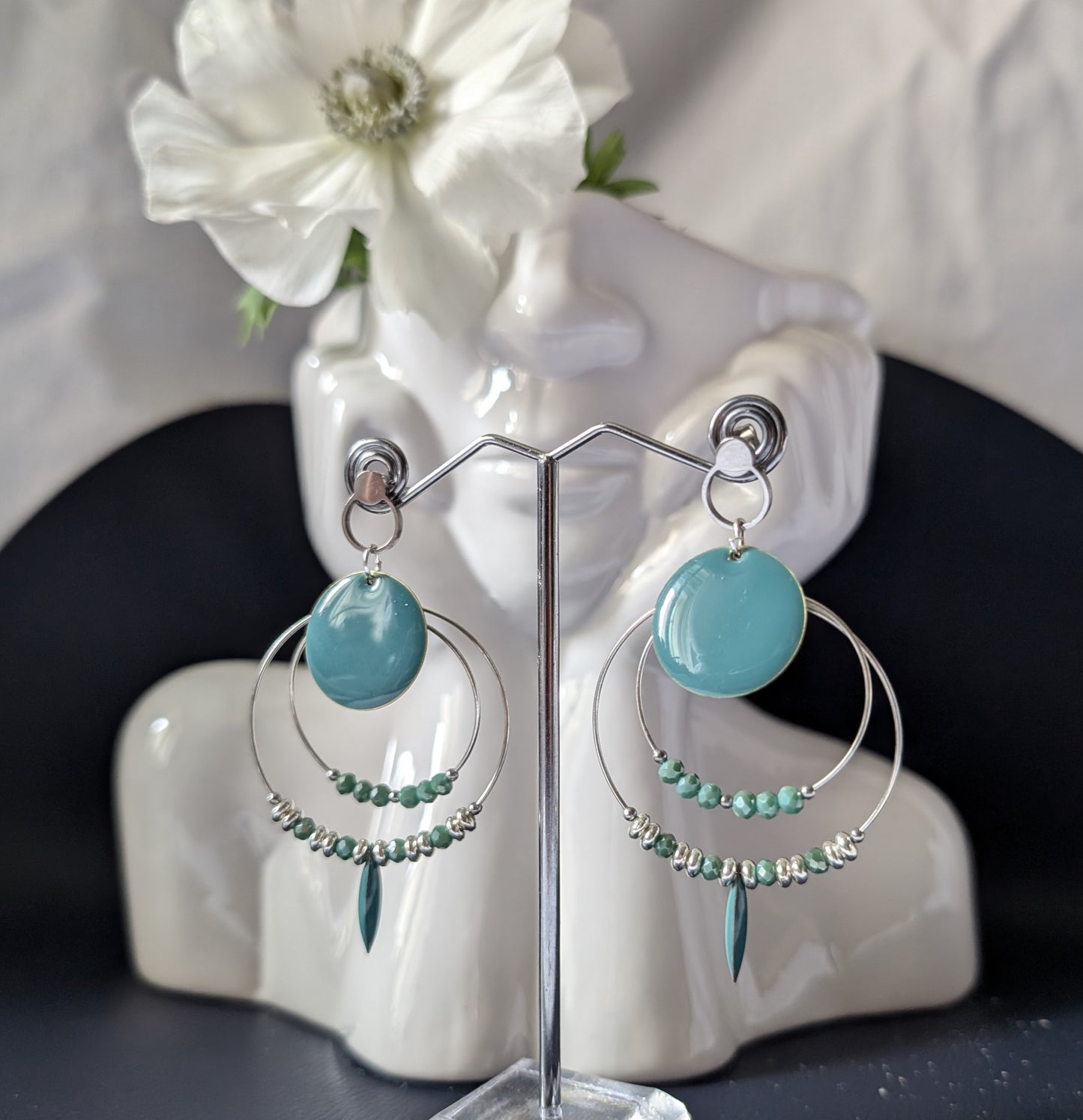 Mise en scene des Boucles d'oreilles Cheyenne devant une vase en forme de buste. Elles sont en acier inoxydable argenté, avec perles de verre couleur jade, perles argentées et éléments en laiton émaillé, sur créoles de tailles variées