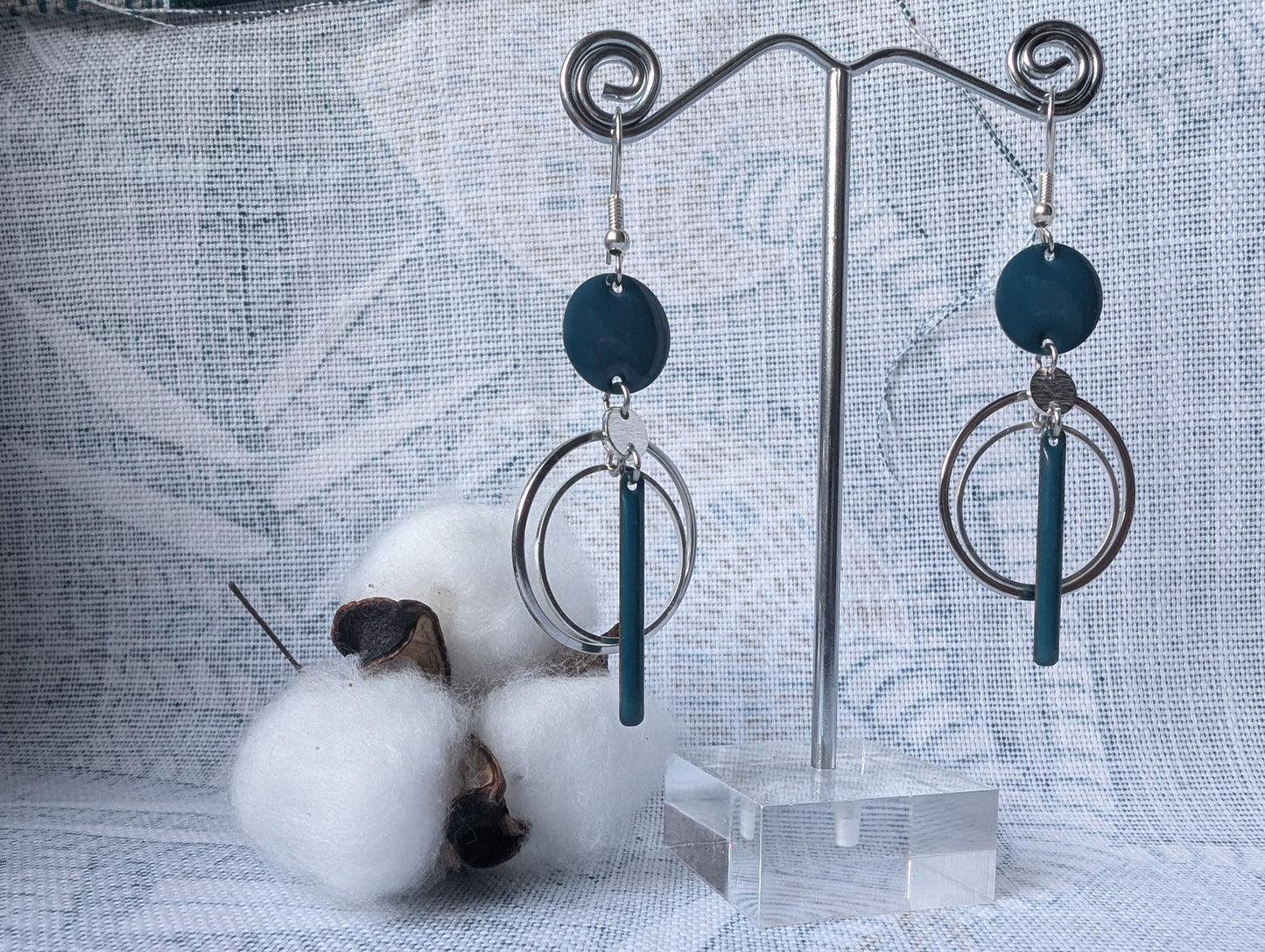 Boucles d’oreilles Infini Bleu en acier inoxydable argenté avec connecteur en laiton émaillé bleu canard, et plusieurs cercles en acier inoxydable, design minimaliste et élégant dans un décor cotonneux.