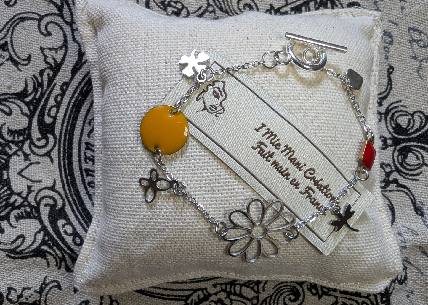 Gros plan sur le bracelet Floréale, mettant en valeur ses motifs floraux éclatants et son fermoir toggle en acier inoxydable. Mise en scène sur petit coussin et marque.