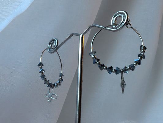 "Vue de profil Boucles d’oreilles Colombie en acier inoxydable, ornées d’un éclat de verre bleu-gris et d'un pendentif papillon. Un bijou élégant et raffiné, aux nuances délicates."