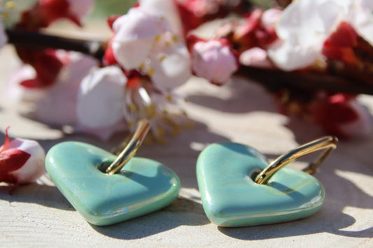 Boucles d’oreilles Petit Cœur de Pierre – Créoles en acier inoxydable doré avec pendentif cœur en céramique turquoise. Bijou délicat et intemporel. Mise en scène fleurs.
