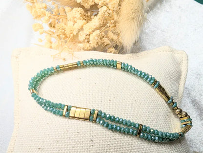 Bracelet Ondine en gros plan, mettant en valeur les perles de verre cyan irisé et les touches dorées d’hématite.Mise en scene petit coussin et fleurs séchées