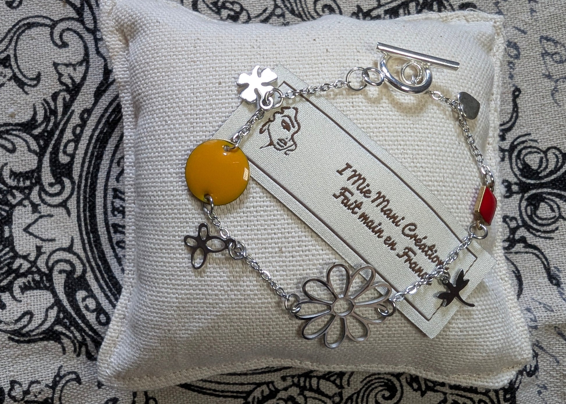 Gros plan sur le bracelet Floréale, mettant en valeur ses motifs floraux éclatants et son fermoir toggle en acier inoxydable. Mise en scène sur petit coussin et marque.
