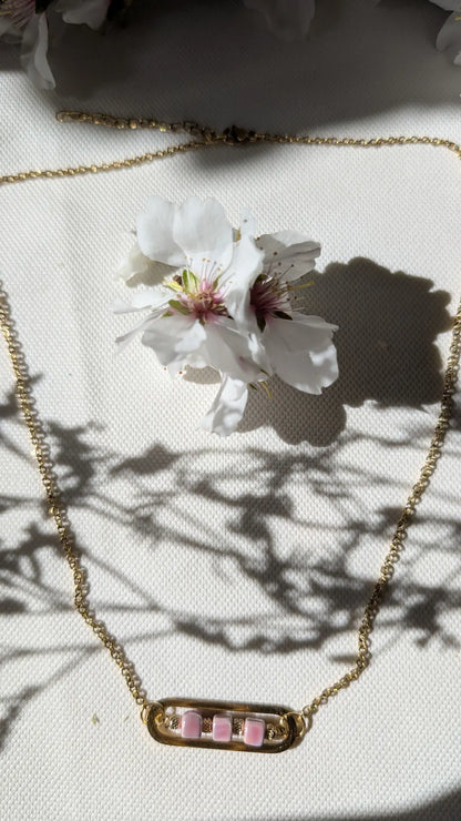 Collier Roséa avec chaîne dorée et perles en céramique rose, disposé en cercle sur un fond neutre pour sublimer son éclat.